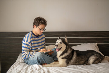Teenage boy and Siberian husky sitting together on bed, company better than any classmate, moment impossible to repeat on second day. Wide shot. Concept of studying together with own dog © iwavephoto