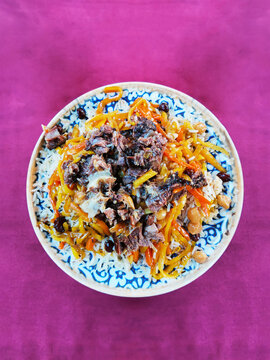 Top-down view of traditional Uzbek plov with meat, rice, carrots, and chickpeas in a ceramic plate on a purple tablecloth. National cuisine dish for restaurant menu and food blog backgrounds.
