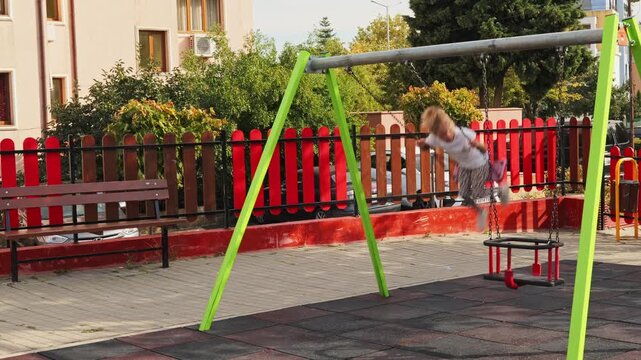 Little girl swinging high on a red swing set at an outdoor playground, enjoying active play on a bright summer day.