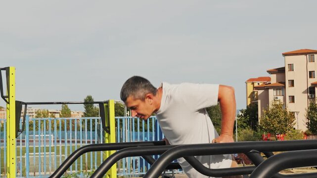Adult man performing dips on parallel workout bars at an outdoor street workout gym area on a bright sunny day.