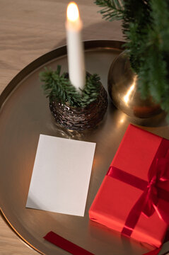 Blank paper card mockup, red gift box, candle and fir branches on golden tray background, festive Christmas holiday still life