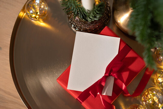 Christmas presents concept, red gift box and blank paper card template on golden tray with garland lights and pine branches, aesthetic holiday background with copy space