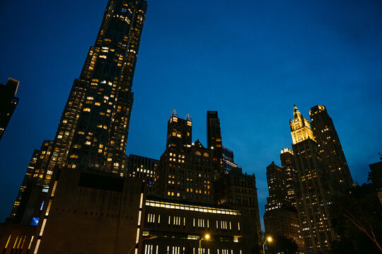 Tall skyscrapers and historic buildings glow with warm lights against the deep blue night sky in Lower Manhattan. The illuminated city skyline highlights the dramatic architecture of New York City.
