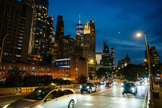 Vehicles move along a busy Manhattan roadway under glowing streetlights and towering skyscrapers. The illuminated skyline captures the vibrant energy of New York City at night.