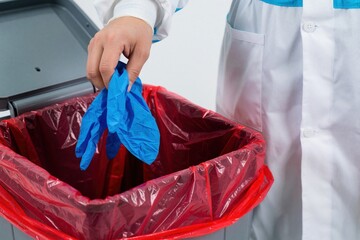 Fototapeta premium Medical staff discard used blue nitrile gloves into red biohazard waste bin. Doctor in white lab coat performs disposal of infectious medical equipment. Proper hygiene, safety protocols for managing