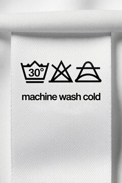 Close up of white fabric laundry label with black care symbols. Text says machine wash cold. Clothing tag shows washing instructions for textile maintenance. Detail of garment care sign on cotton