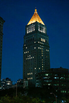 A historic Manhattan skyscraper with a golden pyramid roof shines against the dark night sky. The illuminated building stands among surrounding city architecture in Lower Manhattan.