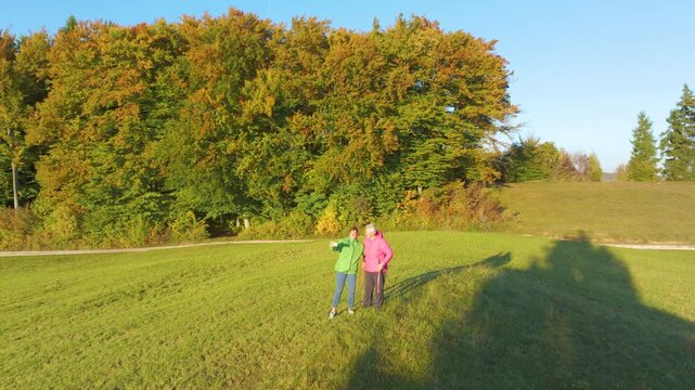 AERIAL: Young and elderly women pause during autumn hike to take a selfie together in sunny meadow. Colourful woodland and warm evening light frame an authentic moment of intergenerational bonding.