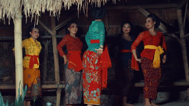 A group of Indonesian women wearing diverse colorful lace Kebayas and Batik skirts practicing traditional dance movements inside a rustic bamboo hut.