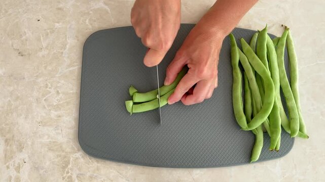 The woman is cutting fresh green pods with a metal knife and preparing seasonal green beans on a gray cutting board. Top‑down view, theme: fresh harvest, healthy vegetables, and home cooking