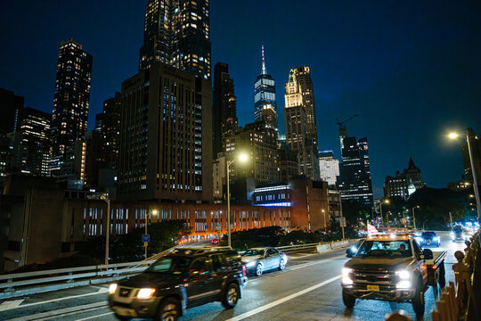 Heavy traffic moves through a Manhattan roadway under glowing streetlights and tall illuminated skyscrapers. The bright city skyline captures the vibrant nightlife of New York City.