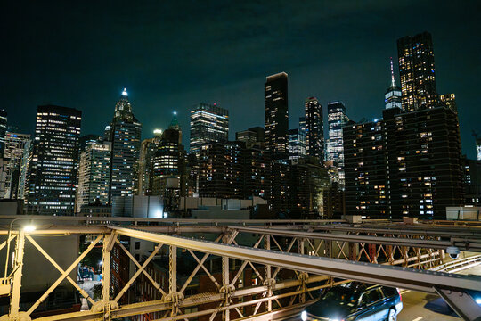 Illuminated skyscrapers of Lower Manhattan rise behind the steel structure of the Brooklyn Bridge at night. Bright city lights and modern towers create a dramatic New York City skyline.