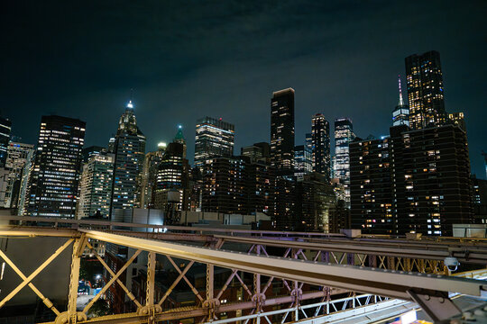 Modern skyscrapers and historic towers glow with lights in Lower Manhattan at night. The steel framework of the Brooklyn Bridge crosses the foreground beneath the illuminated skyline.