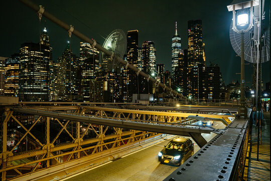 Cars drive across the illuminated Brooklyn Bridge at night with the bright Manhattan skyline in the background. Skyscrapers and city lights create a dramatic urban night cityscape in New York City.