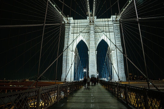 People walk along the wooden pedestrian walkway of the Brooklyn Bridge at night in New York City. The illuminated bridge tower and suspension cables create a dramatic perspective of the historic