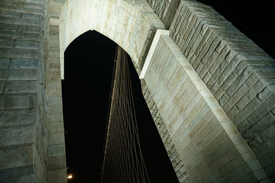 Low angle view of the historic Brooklyn Bridge stone arch and suspension cables against the dark night sky in New York City. Dramatic architectural perspective highlighting the massive stone structure