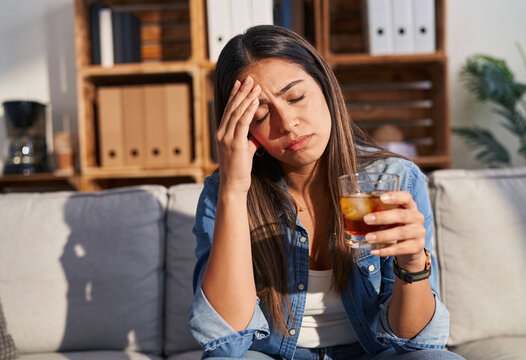 Distressed Hispanic woman holding whiskey glass with hand on forehead sitting on living room sofa coping with stress