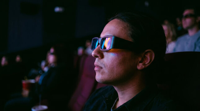 A young man wearing glasses is watching a movie in a dark theater.
