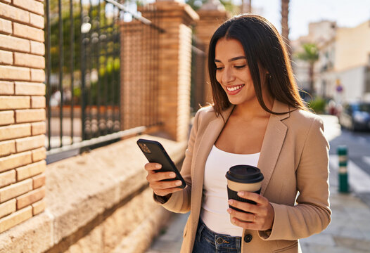 Smiling young hispanic woman using smartphone holding coffee cup on sunny urban street wearing beige blazer and jeans