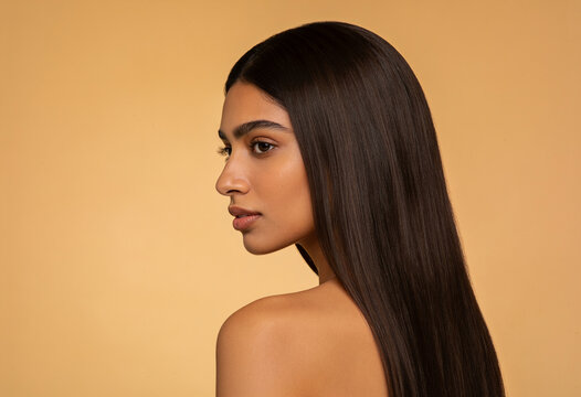 Young South Asian woman with long straight dark hair in side profile posing against warm orange studio background