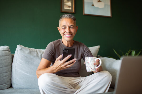 Smiling older woman using smartphone with mug at home