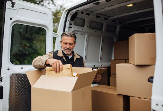 Mature delivery driver checking cardboard box package in white van outdoors natural light logistics courier service