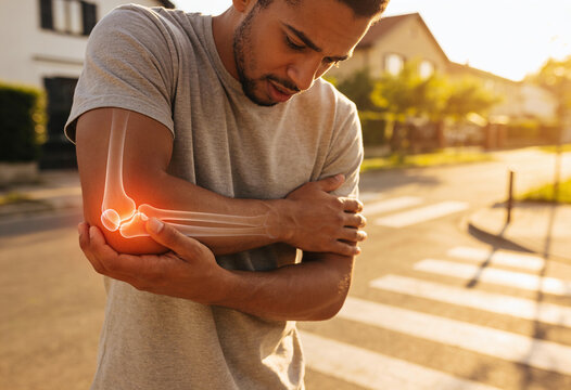 Pained African American man clutching elbow with glowing anatomical bone overlay on sunny suburban street at sunset