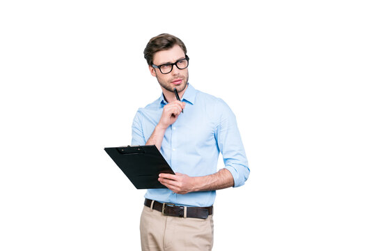 Portrait  of attractive, clever, stunning man in shirt, pants and glasses touching chin with pen, holding clipboard, looking to the side with thoughtful expression over grey background