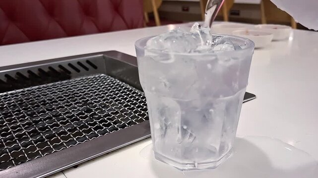 Pouring Fresh Cold Water Into Clear Plastic Glass Filled With Ice Cubes On Table At Korean Barbecue Restaurant
