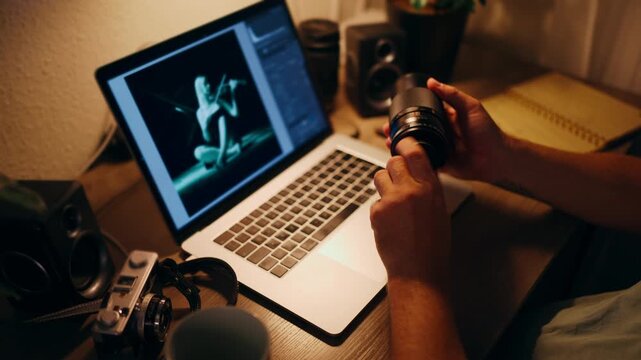 Creative professional photographer examining a camera lens at a warmly lit desk, with a laptop displaying a photo editing software interface and a vintage film camera nearby