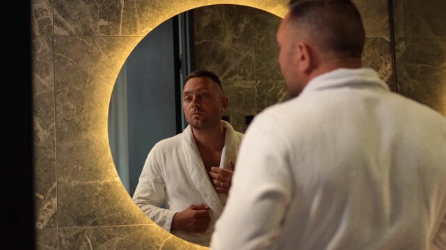 Handsome man in a white bathrobe shaving his face with a razor looking in a round illuminated mirror with a static camera shot.