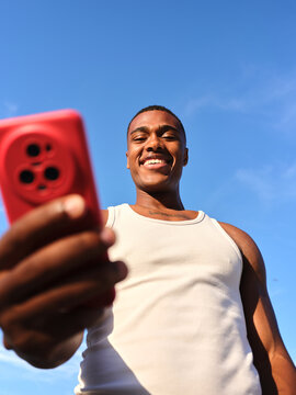 Young man holding red smartphone smiling against blue sky
