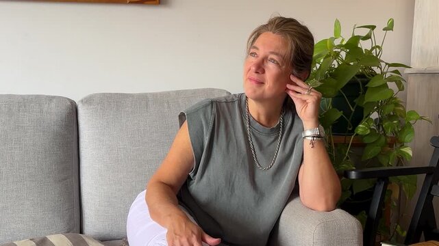 A mature woman who is pensive and relaxed is portrayed as sitting on the couch in her living room.