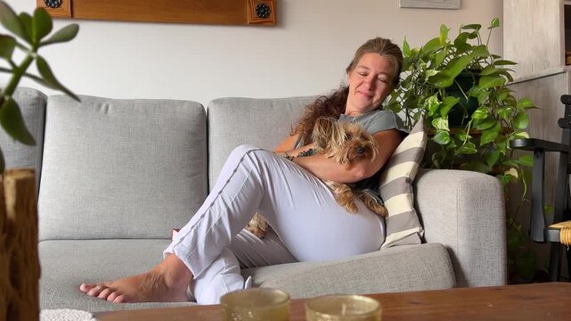Mature woman sitting on a sofa in a modern living room, smiling and petting her cute dog. Concept of serenity and peace of mind