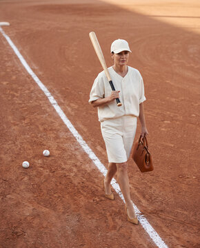 Woman in a retro-style white sports outfit and cap standing on a clay field holding a bat, with balls at her feet. Vintage sporting and lifestyle fashion concept.