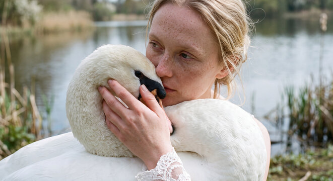 Woman wearing a plush white faux fur coat gently holding a small white bird near a calm body of water. Dreamy winter fashion and nature concept.