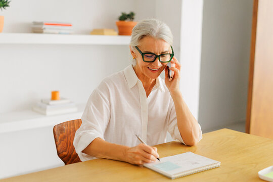 Senior woman teleworking communicating by phone writing notes