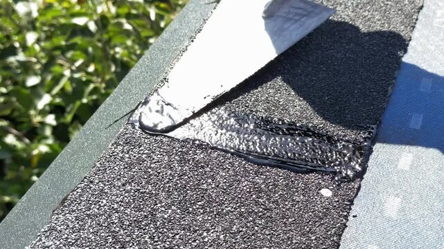 Worker applying bitumen mastic to asphalt shingle seam, roof waterproofing repair