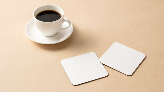 White coffee cup with black coffee and two blank square coasters on a beige background