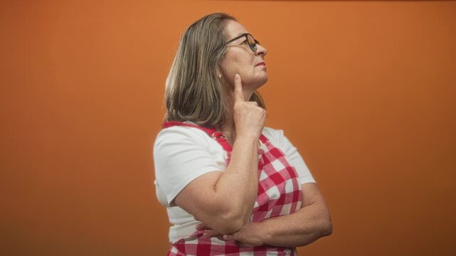Hispanic woman in gingham apron, profile with arms crossed, touching cheek with finger in studio orange backdrop; thoughtful reflection.