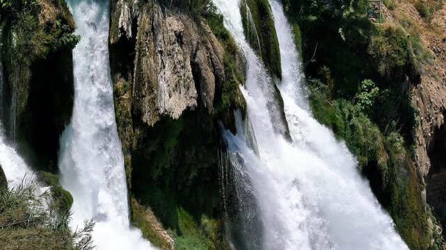 A beautiful waterfall cascades down rugged, moss-covered rock formations, surrounded by lush green vegetation.