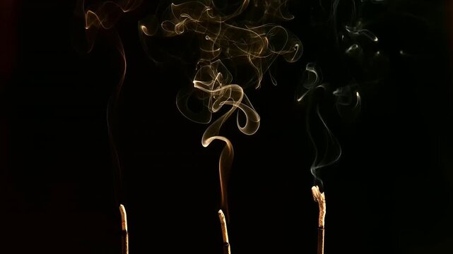 Close-up of three burning incense sticks releasing delicate curling smoke against a deep black background, thin aromatic trails forming abstract shapes in darkness, Antyesti, Antim