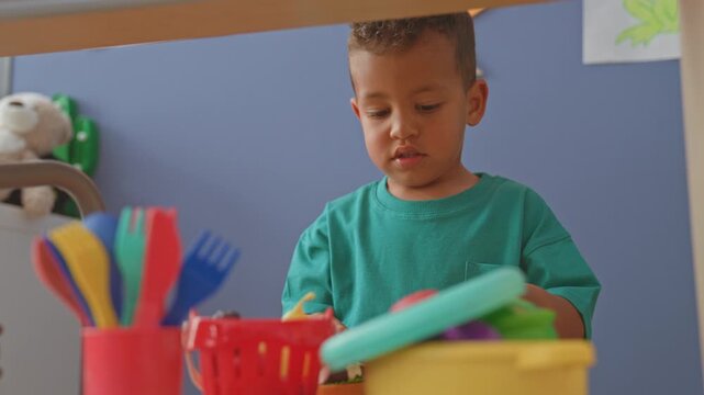 Boy holding a toy burger and playing with plastic utensils at a playroom table; quiet concentration.