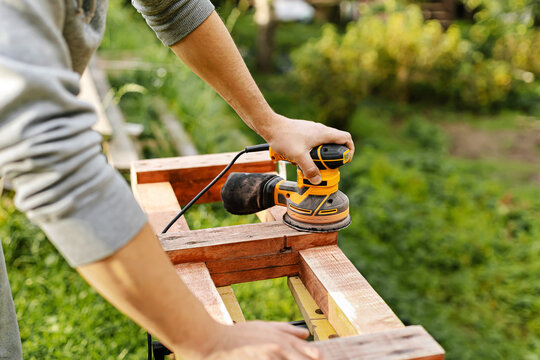 Man sanding wooden furniture with electric orbital sander outdoors. DIY home renovation, carpentry lifestyle concept.