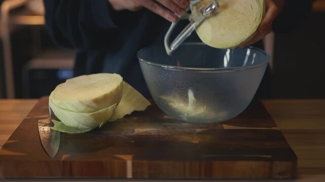 Close-up of woman's hands shredding fresh cabbage with a peeler into a glass bowl on a wooden board on a table. Cooking and healthy food preparation concept in a cozy warm home kitchen.