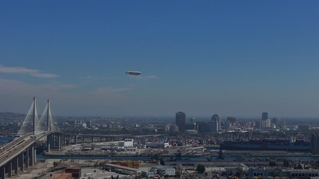 Aerial view of the Gerald Desmond Bridge and the bustling Port of Long Beach with a blimp flying over the harbor under a clear blue sky in Long Beach, California, United States.
