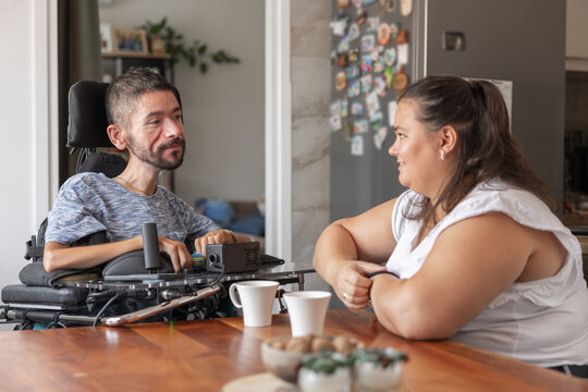 A man in a wheelchair talks with a woman in a kitchen. They share a moment over cups and snacks, showing support and friendship.