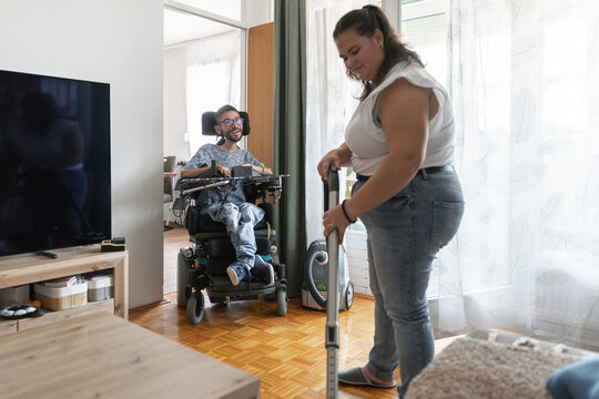 A woman vacuuming floor the floor with a man in a wheelchair in their home. They are engaged in daily living activities together.