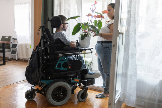 A man in a wheelchair sits in a room while a woman stands nearby holding and watering a plant. They are engaging in conversation in a home setting.