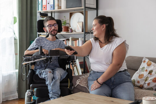 A man in a wheelchair sits beside a woman in a living room while she hands him the remote control. They both smile and watch TV.